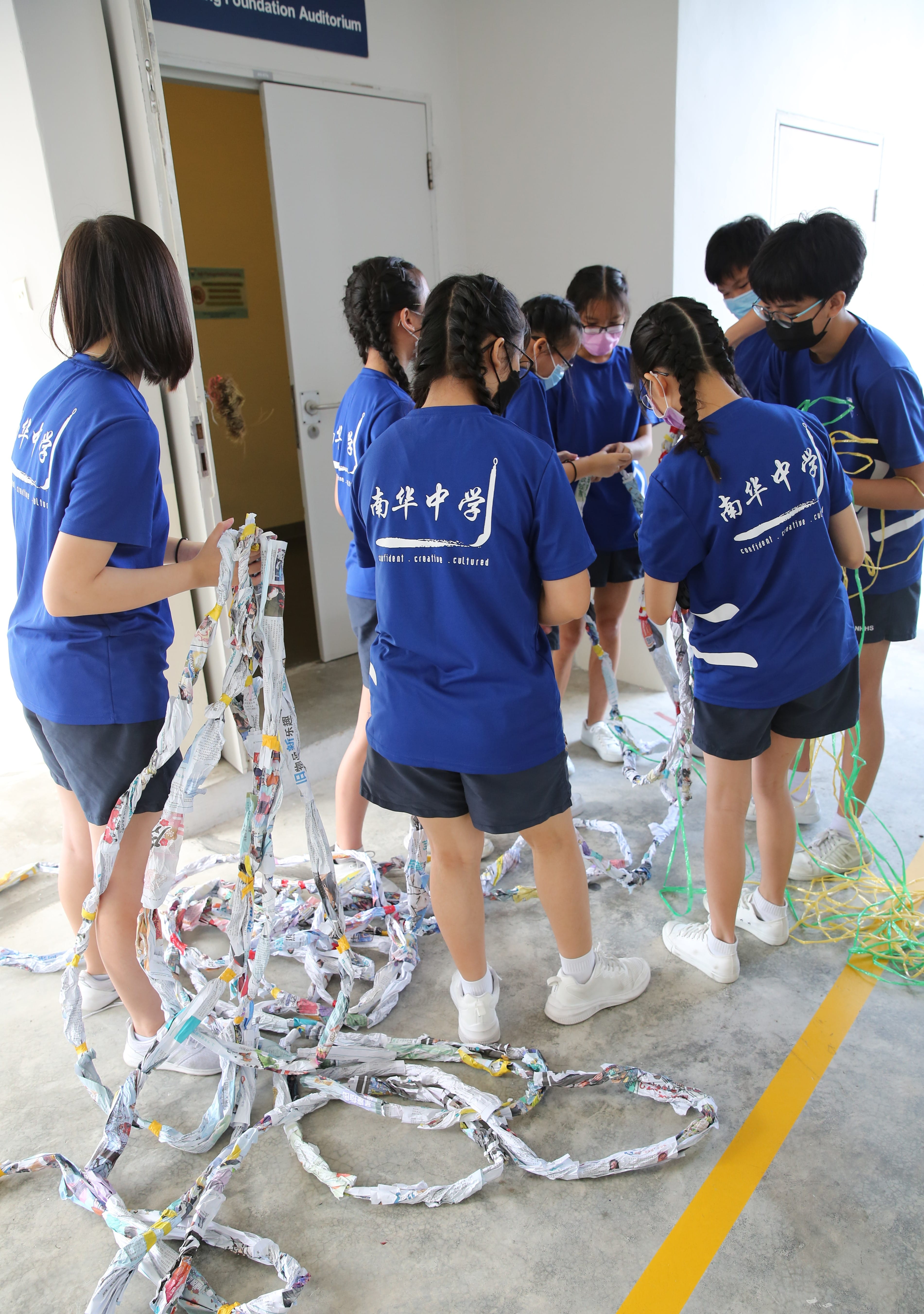 Students in blue uniforms work together weaving twisted newspaper strips and colorful cords on a concrete floor.